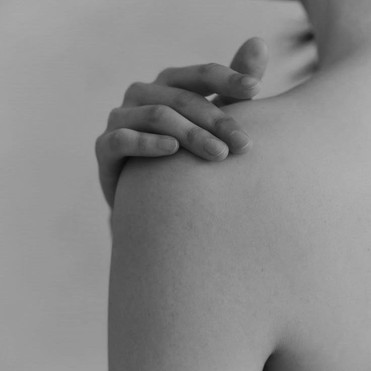 Close up black and white photo of a hand resting gently on a bare shoulder, highlighting smooth skin and a calm, minimal aesthetic often associated with cosmetic and skin treatments.