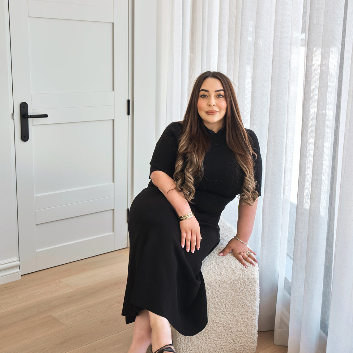 A woman in a fitted black dress sits by a sunlit window with sheer curtains inside a modern cosmetic clinic, presenting a polished and welcoming atmosphere for aesthetic treatments.