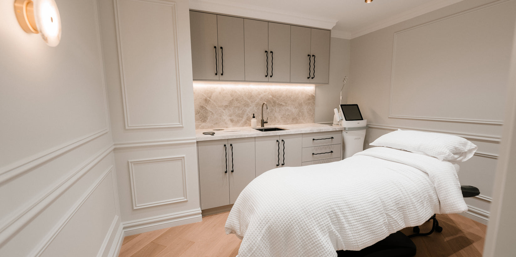 Elegant cosmetic treatment room with a padded white treatment bed, soft ambient lighting, and built in cabinetry with a sink, reflecting a clean and calming environment for injectable cosmetic procedures.