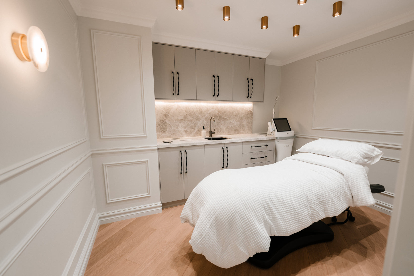 Elegant cosmetic treatment room with a padded white treatment bed, soft ambient lighting, and built in cabinetry with a sink, reflecting a clean and calming environment for injectable cosmetic procedures.