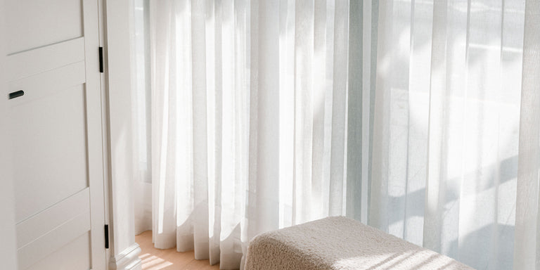 White curtain filtering sunlight in a room with a textured bench on a wooden floor.